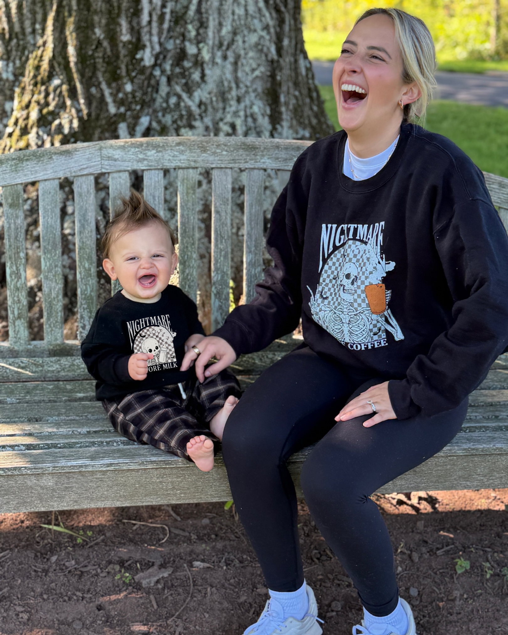 A woman and a young child sitting on a wooden park bench, both laughing and happy. They are outdoors with trees and grass in the background, dressed casually in black sweatshirts with matching skull and ice cream graphic designs.