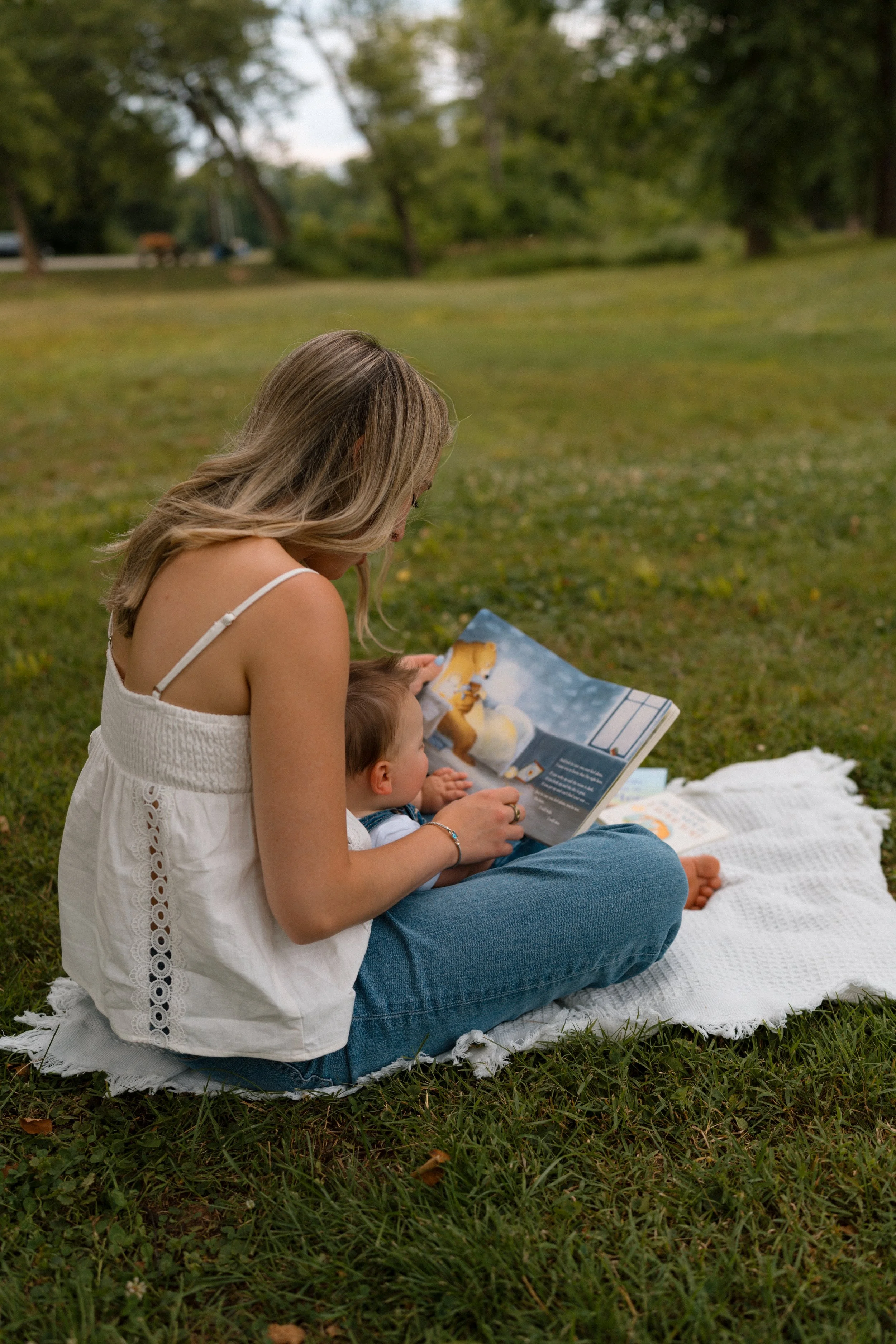 A woman and a young boy sitting on a white blanket on grass, reading a book together outdoors in a park with trees in the background.