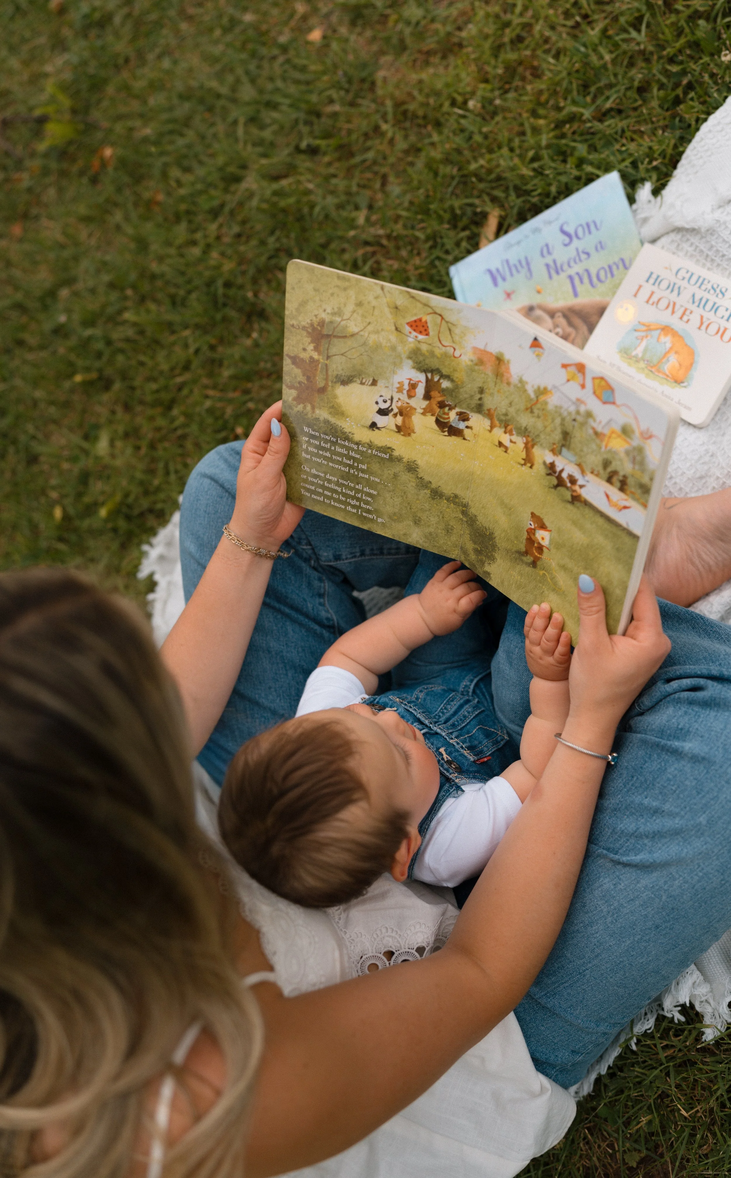 A woman and two children sitting on grass, reading children's books together, with books showing colorful illustrations and titles about family and love.
