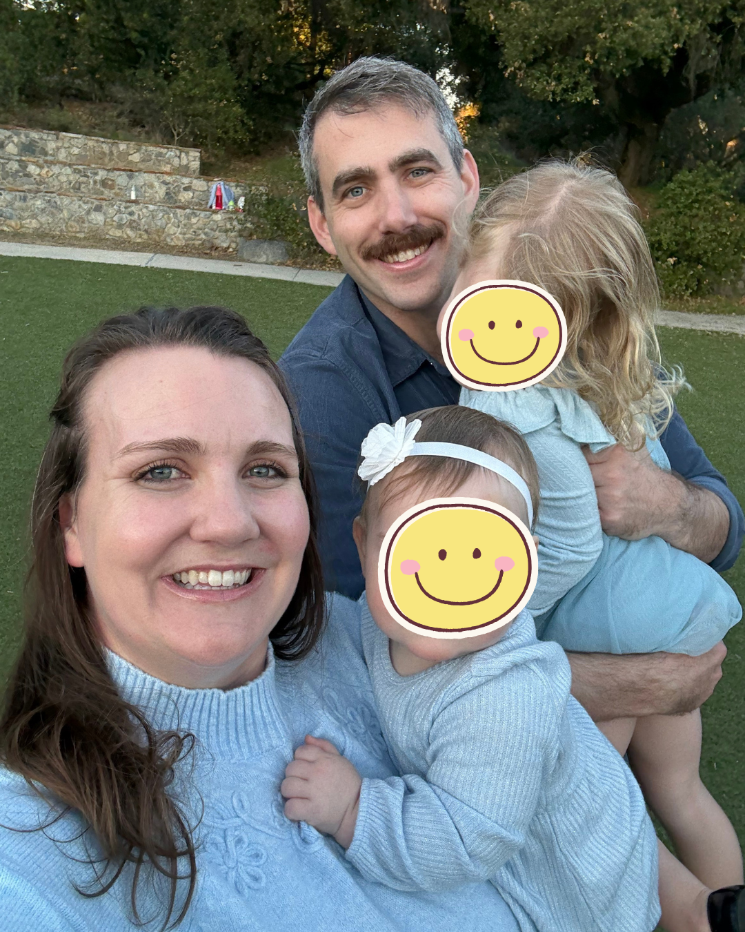 A smiling family of four taking a selfie outdoors on a grassy area with trees in the background. The father is holding the older daughter, and the mother is holding the younger daughter. Both daughters have smiley face stickers over their faces.