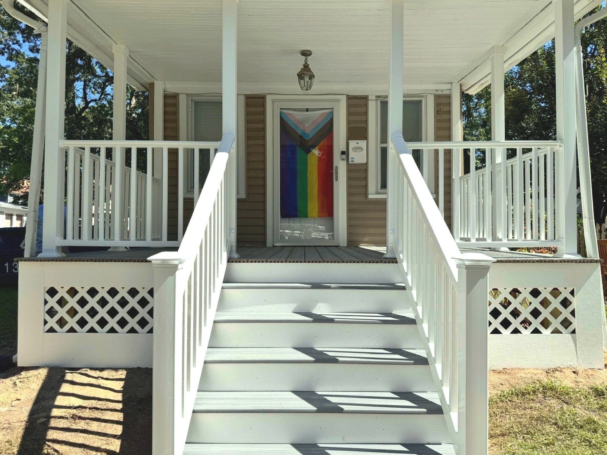 White porch with stairs, railing, and lattice skirting, featuring a rainbow pride flag hanging on the front door, in a sunny outdoor area surrounded by trees.