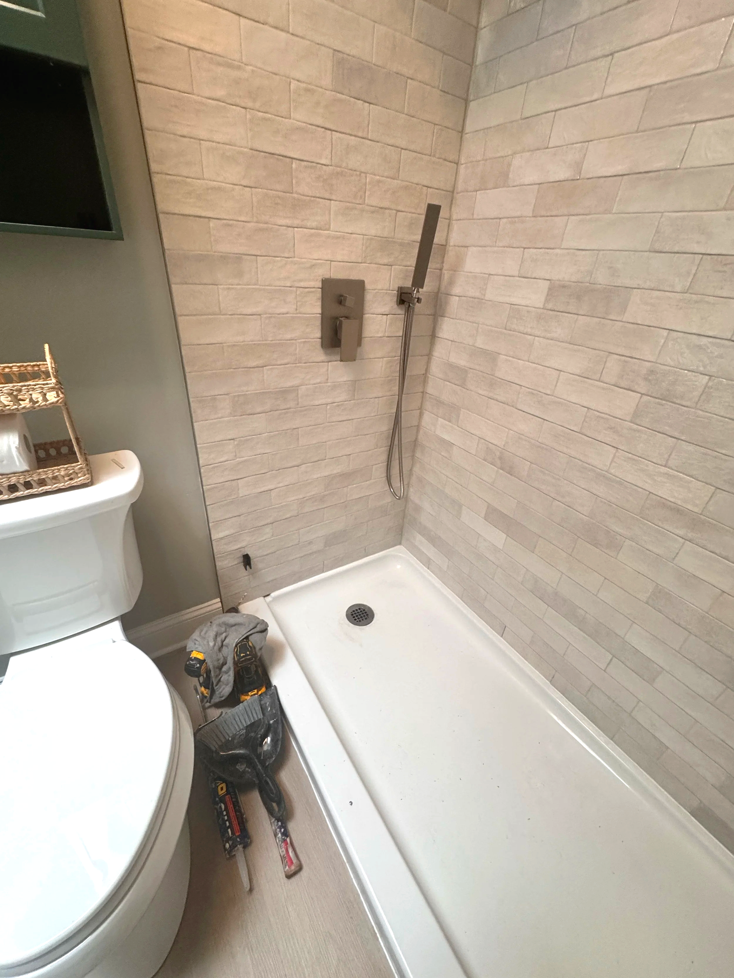 A bathroom shower area with beige tile walls, a shower head, and a drain on the white shower floor. There are tools and a gray cloth on the floor, with part of a toilet and a small storage shelf visible nearby.