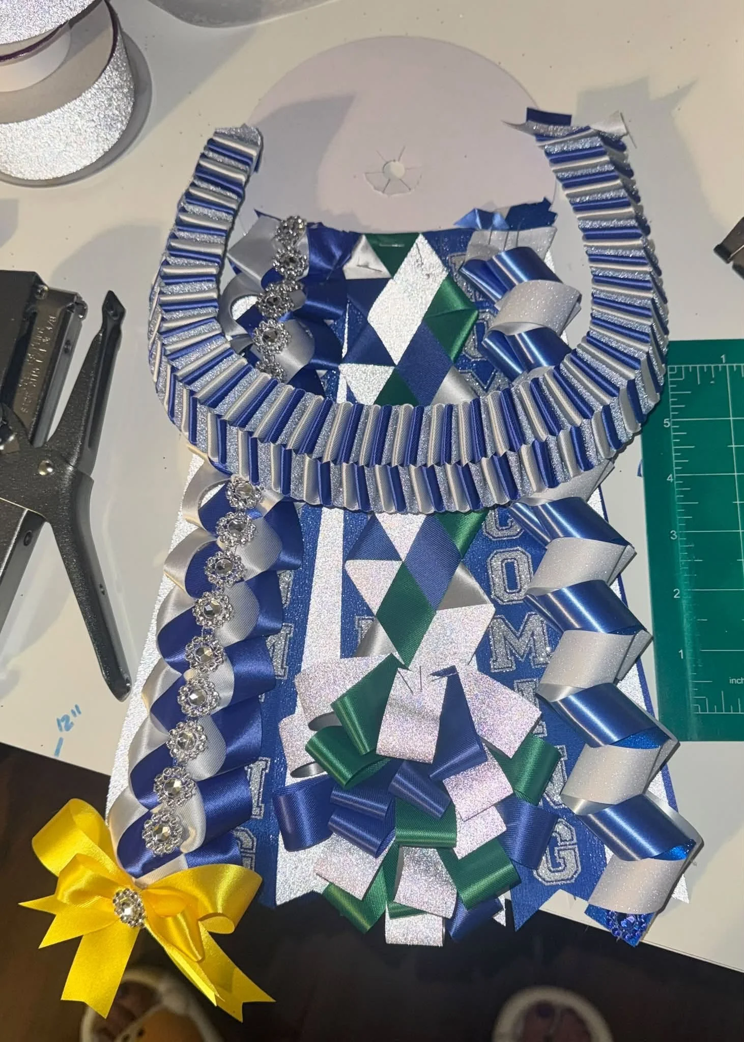 A collection of ribbons and bows in blue, green, yellow, silver, and white with decorative rhinestones, arranged on a white glittery surface, likely for a celebration or event decoration.