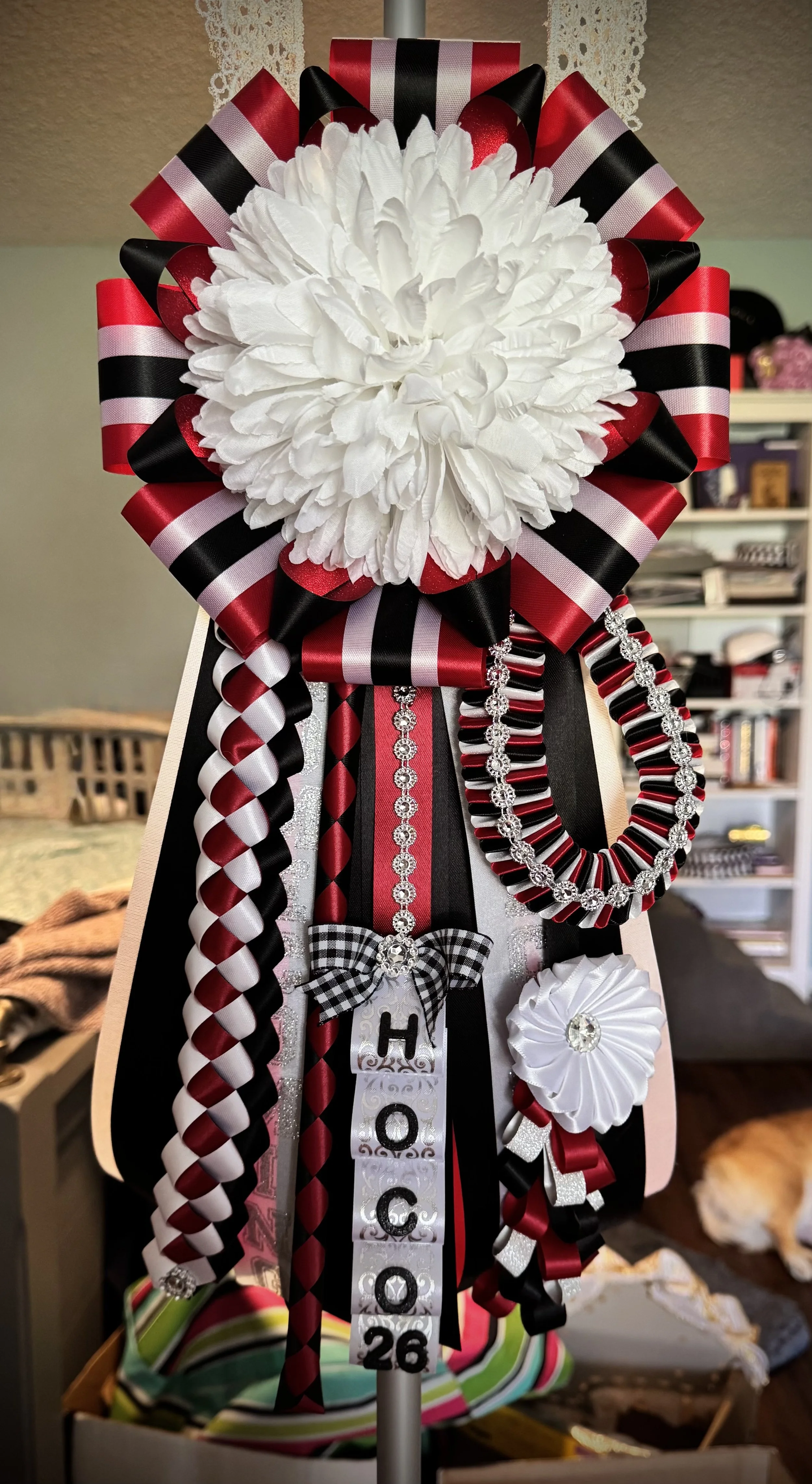 A decorative graduation wreath with a large white flower at the center, surrounded by black, red, and white ribbons and bows, and adorned with silver and rhinestone accents, including a hanging tag with the year '2026' and the word 'Hood'.