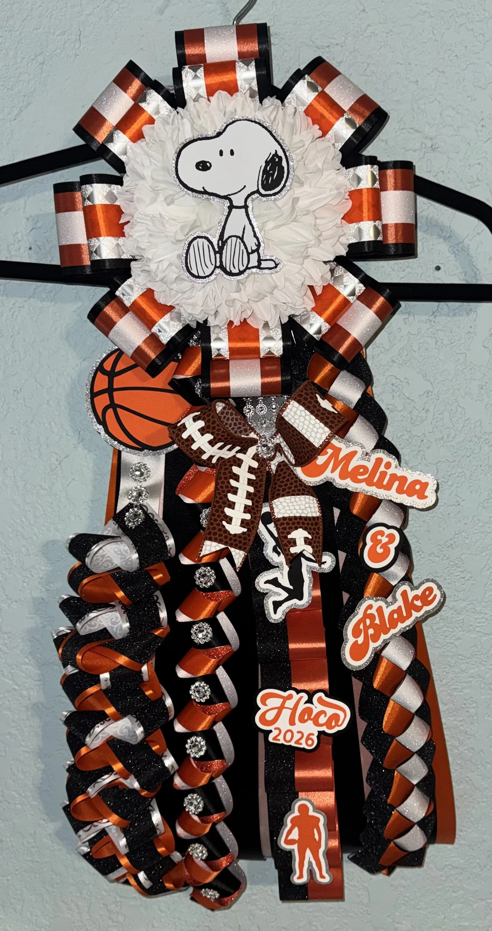 Decorative graduation sash in black, orange, and white with ribbons, rhinestones, and Peanuts character Snoopy applique, with the names Melina, Blake, and a 2026 graduation year sticker.