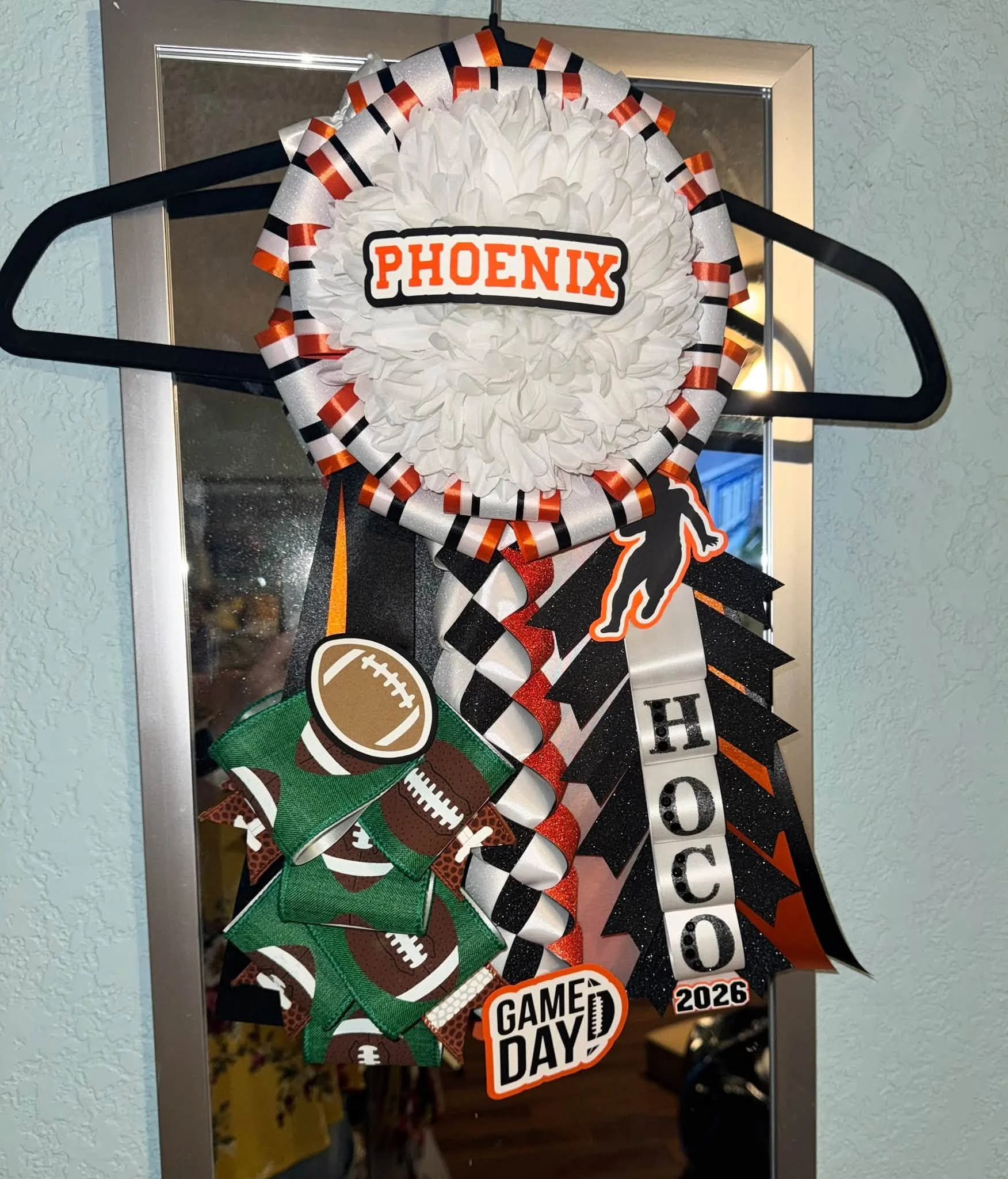 Decorative wreath with the word 'PHOENIX' in the center, surrounded by sports-themed embellishments, ribbons in black, orange, and white colors, and a 'GAME DAY' badge, hanging on a mirror.
