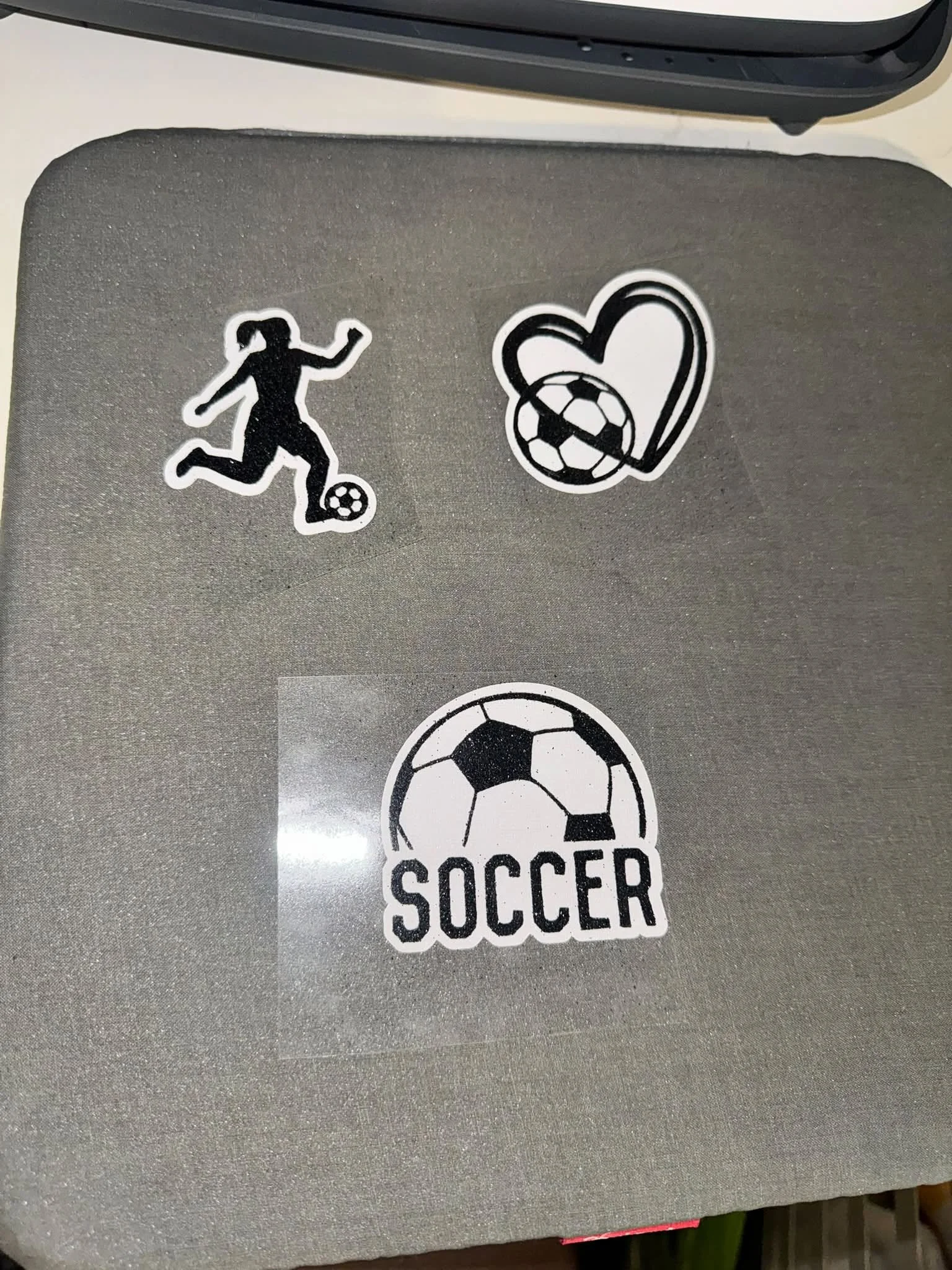 Three soccer-themed stickers on a gray surface: a silhouette of a girl running with a soccer ball, a heart with a soccer ball inside, and a circular logo with a soccer ball and the words "SOCCER".