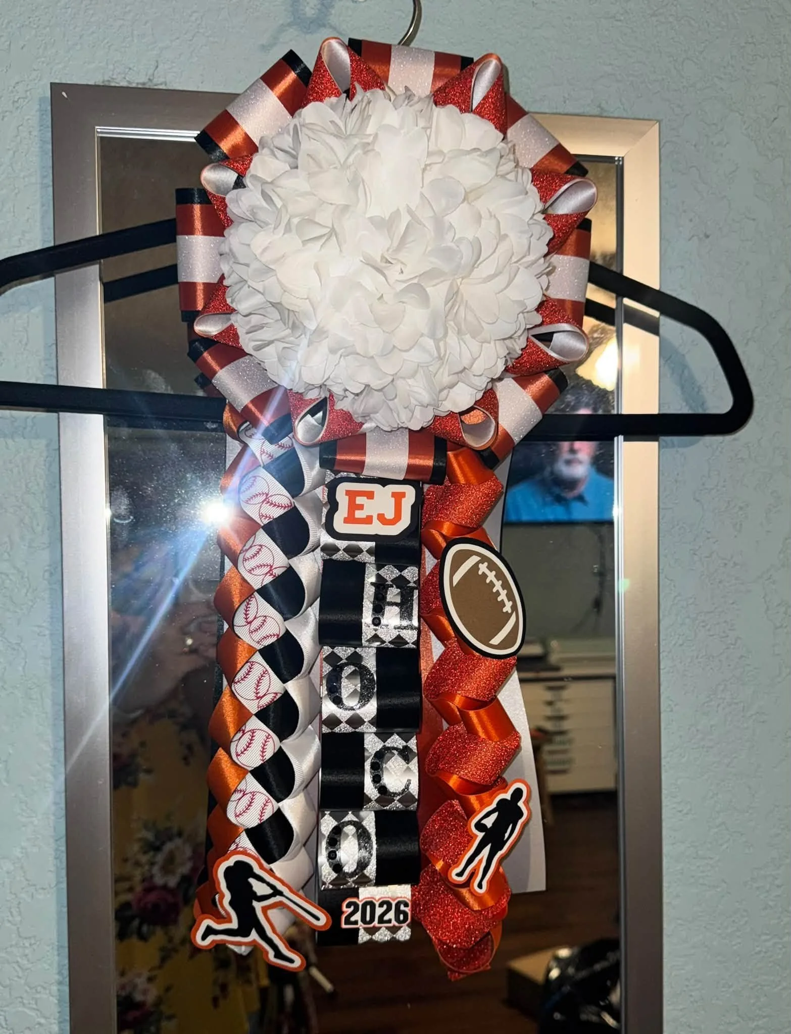 A decorative sports-themed ornament with a white flower at the center, surrounded by ribbons in orange, black, white, and baseball-patterned designs, featuring football and running figure stickers, and the year 2026.