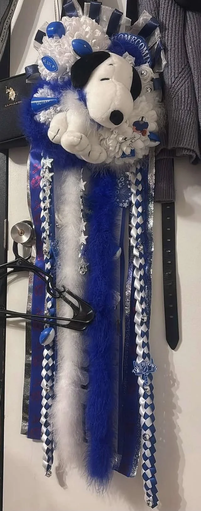 Decorated plush Snoopy dog with blue and white themed ribbons, feather boa, and sports-themed accessories, likely for a celebration or game day, on a table with miscellaneous items.