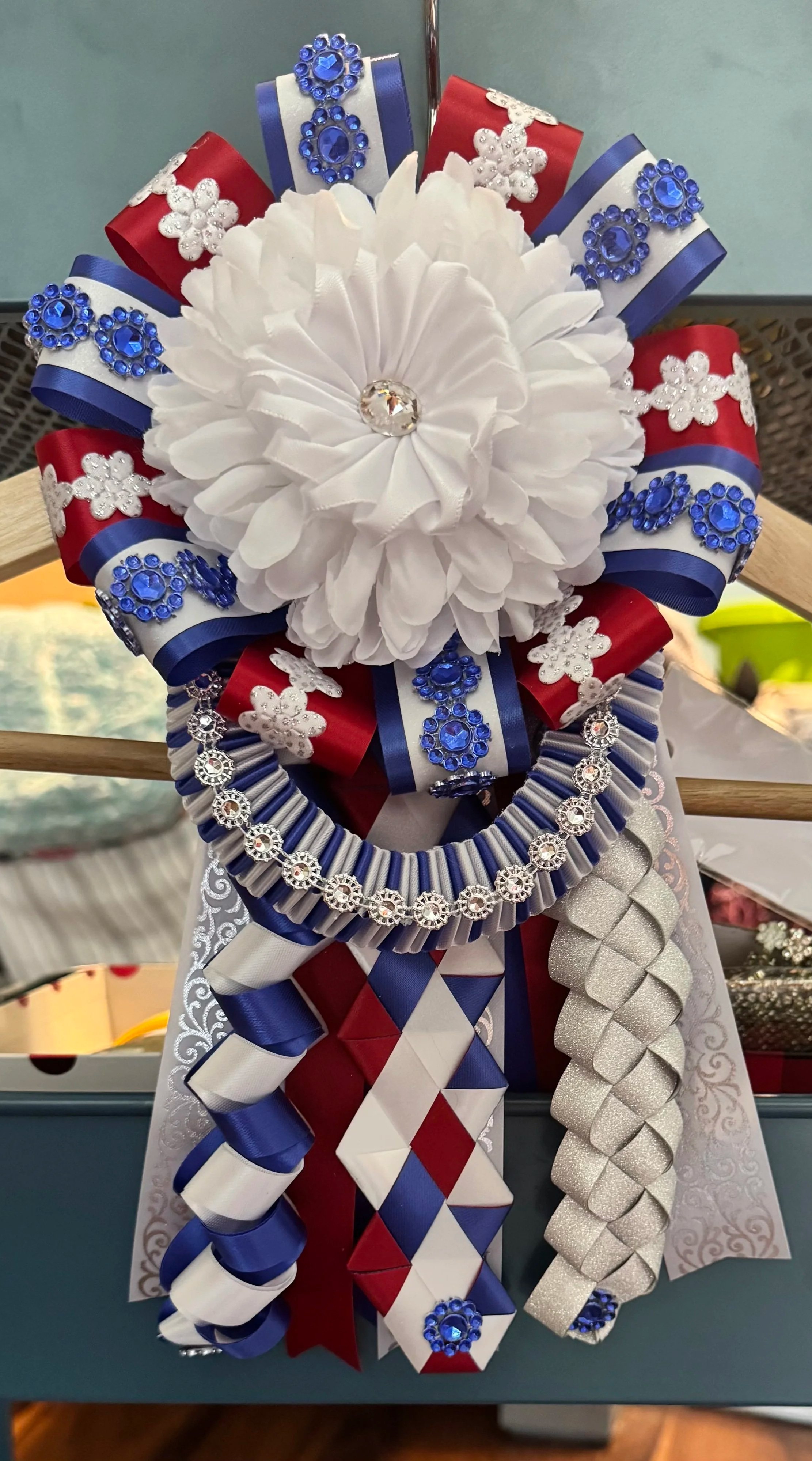 A decorative bow or wreath made with red, white, and blue ribbons, adorned with blue rhinestones, white snowflake embellishments, and a layered white flower at the center, decorated with a large central rhinestone.