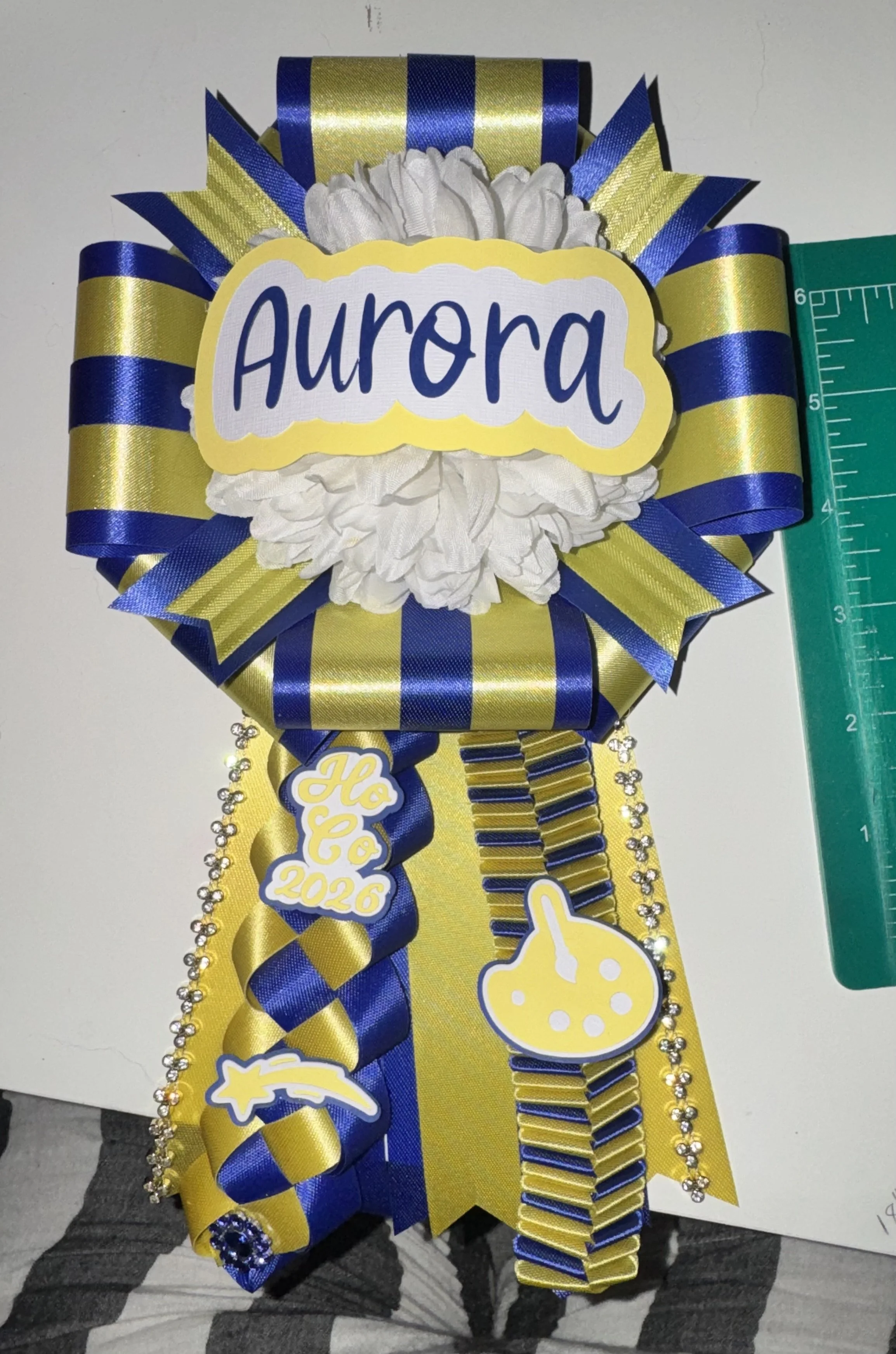 A decorated graduation sash with blue and yellow ribbons, large white flower, and yellow and blue signs saying 'Aurora', 'Hoco 2026', a star and a lollipop, hanging on a person's neck. Green ruler on the side for scale.