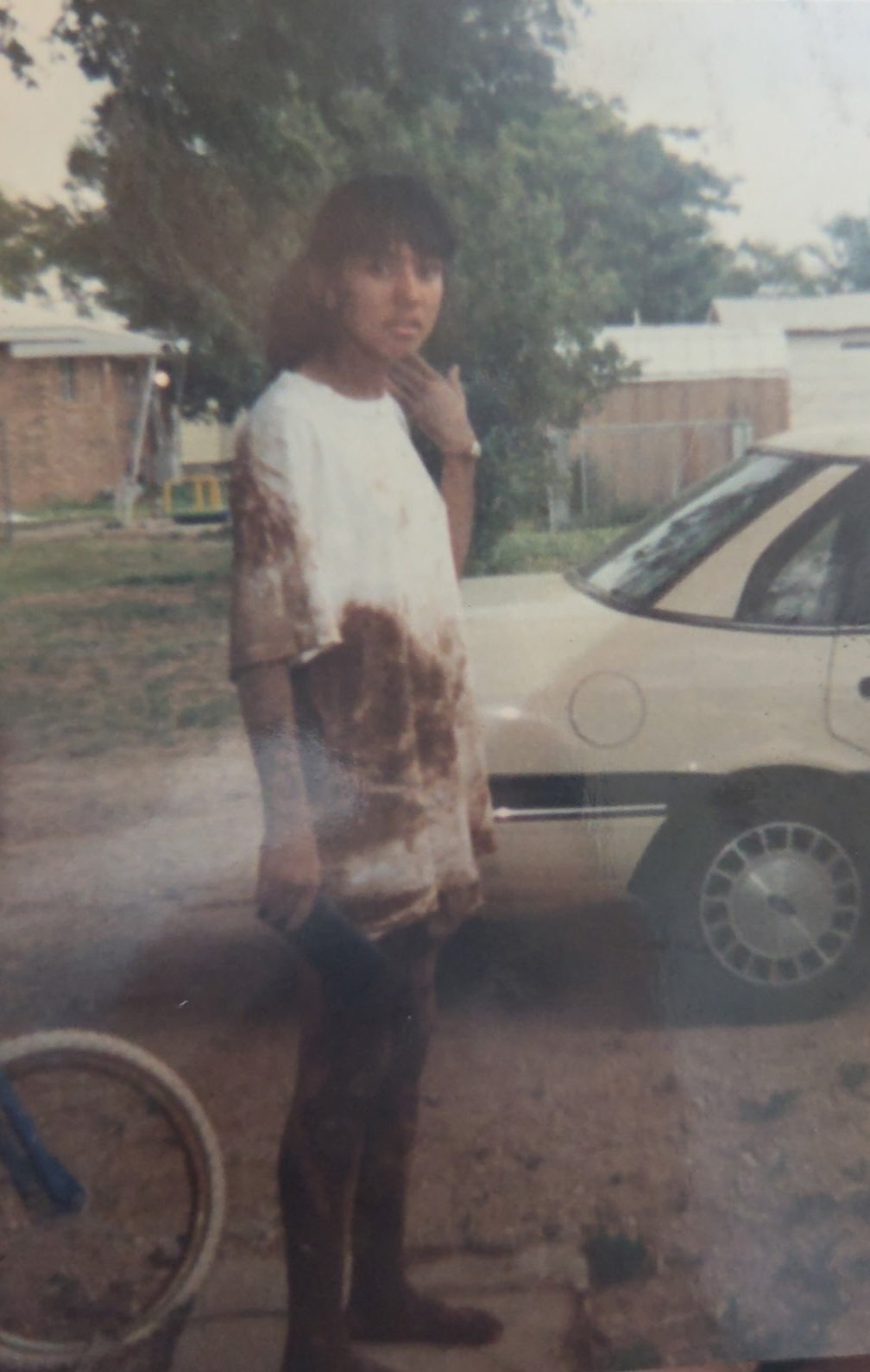 A young woman with short curly hair standing outdoors by a parked car, with a tree and residential buildings in the background.