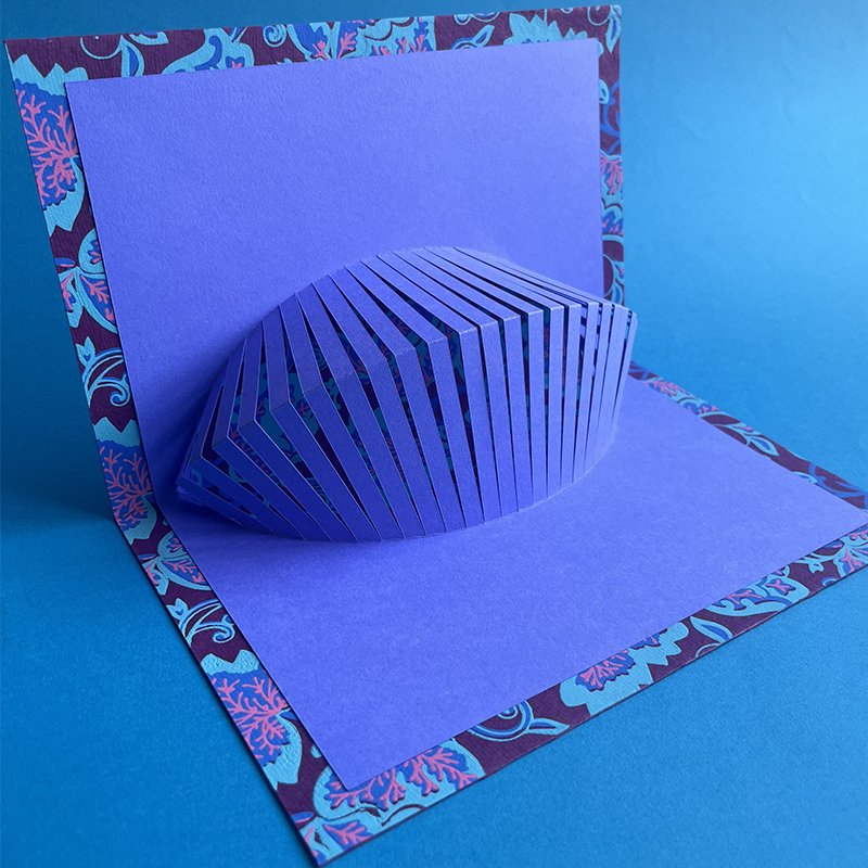 A 3D paper art piece of a purple clam shell, made from layered paper, set on a purple and patterned background with a floral border, all on a blue surface.