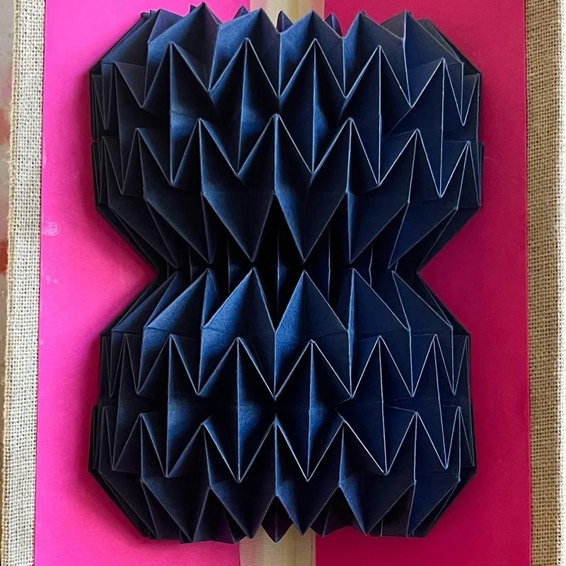 Black accordion-style paper sculpture mounted on a pink background.