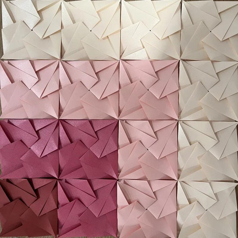 Pattern of folded paper squares in shades of pink, arranged in a grid pattern.
