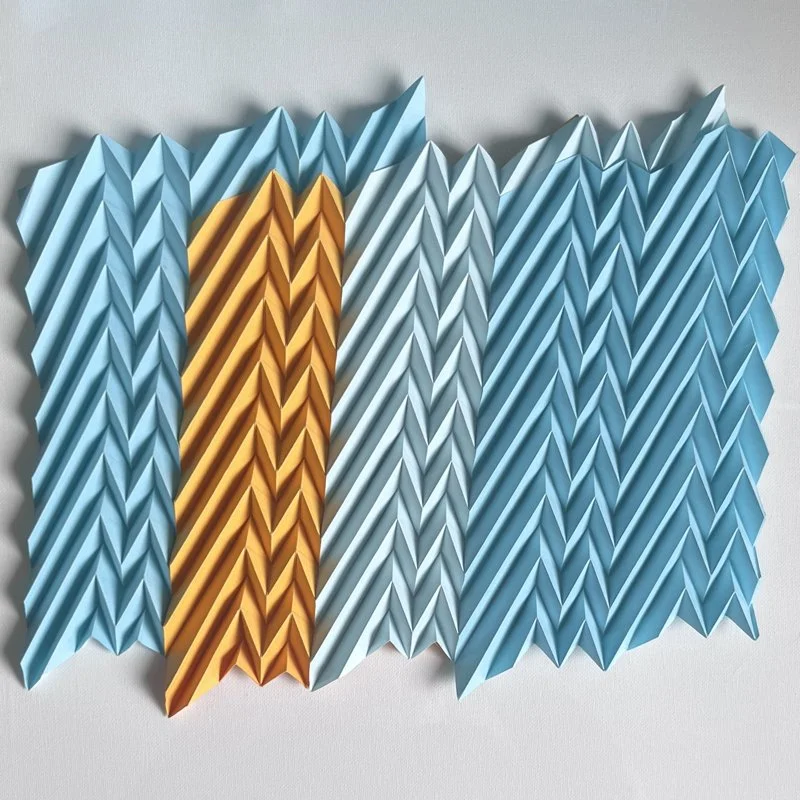 Colorful paper zig zags arranged in a pattern, mainly in shades of blue with one section in orange, creating a dynamic visual effect.