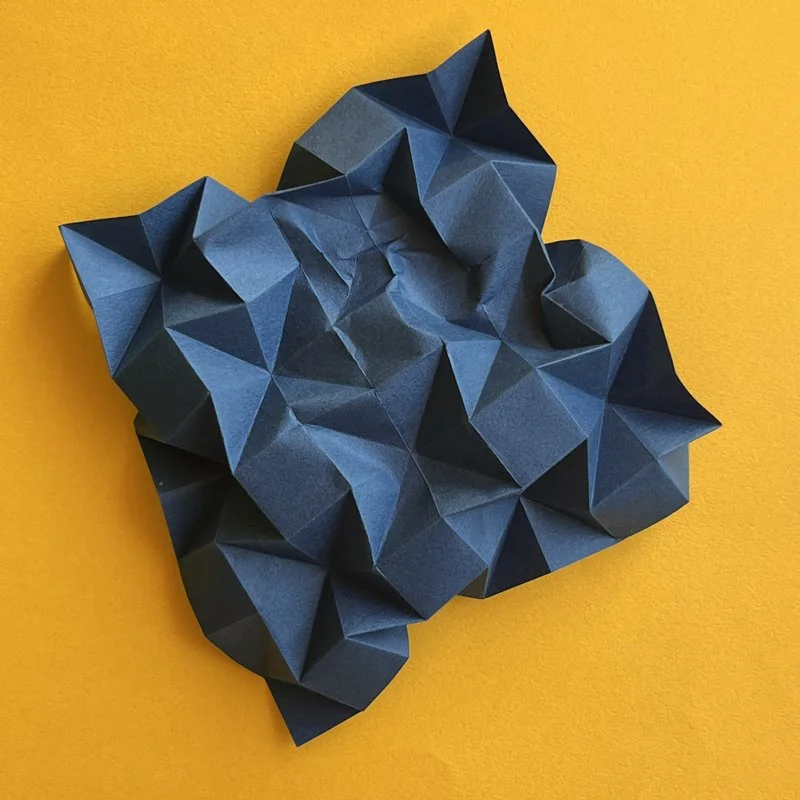 A crumpled, geometric paper sculpture on a yellow background.