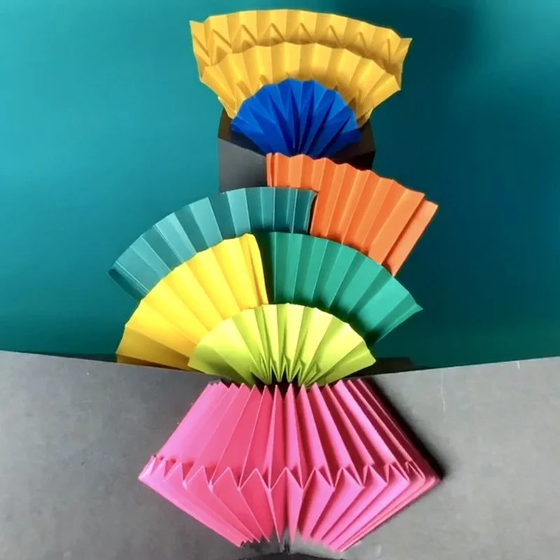 Colorful paper fans arranged in a semi-circle with a pink paper fan at the bottom, forming a rainbow-like pattern against a teal background.