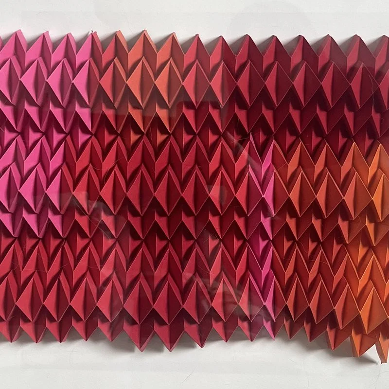 A close-up view of an artistic wall installation made from numerous folded paper shapes in shades of pink, red, and orange, arranged in a geometric pattern.