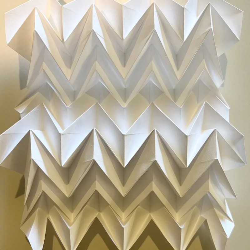 An art installation made of white paper folded into zigzag patterns, arranged in multiple rows on a wall.