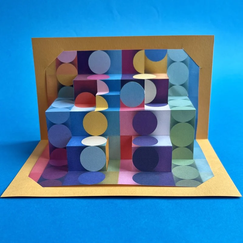 Colorful pop-up with layered cubes and circles creating a geometric pattern against a blue background.