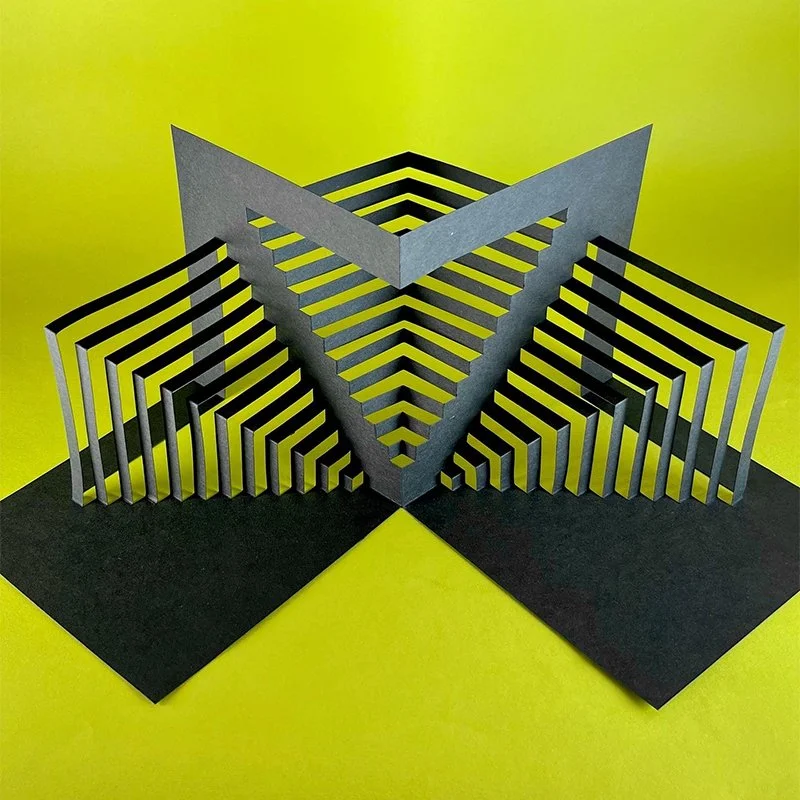 Black and gray geometric optical illusion sculpture with layered angular shapes on a yellow background.