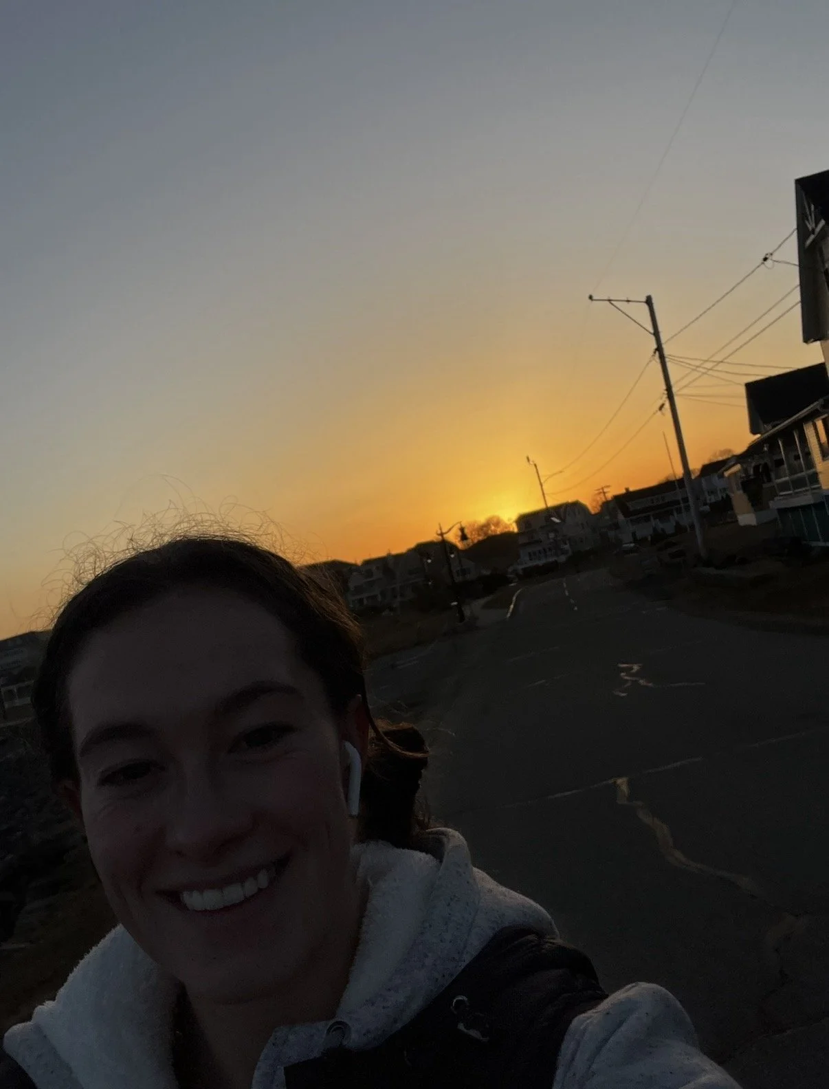 A woman smiling at sunset, with her face partially in shadow. The sky is clear with a warm orange glow near the horizon, and there are buildings and power lines in the background.