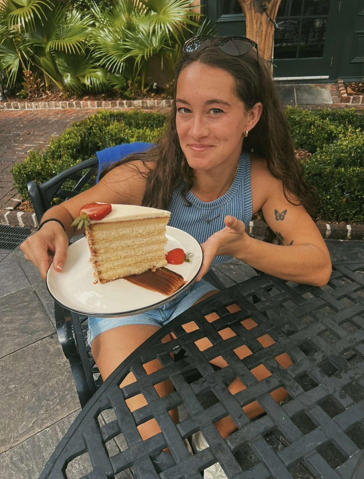A woman sitting outdoors at a dining table, holding a plate with a slice of layered cake topped with a strawberry, with another strawberry on the plate. She is smiling and wearing a blue sleeveless top and sunglasses on her head.