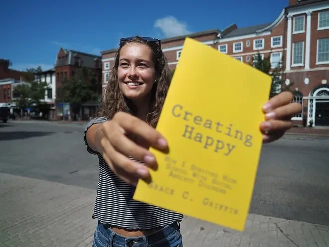 Young woman smiling and holding a yellow book titled 'Creating Happy' outside on a city street.