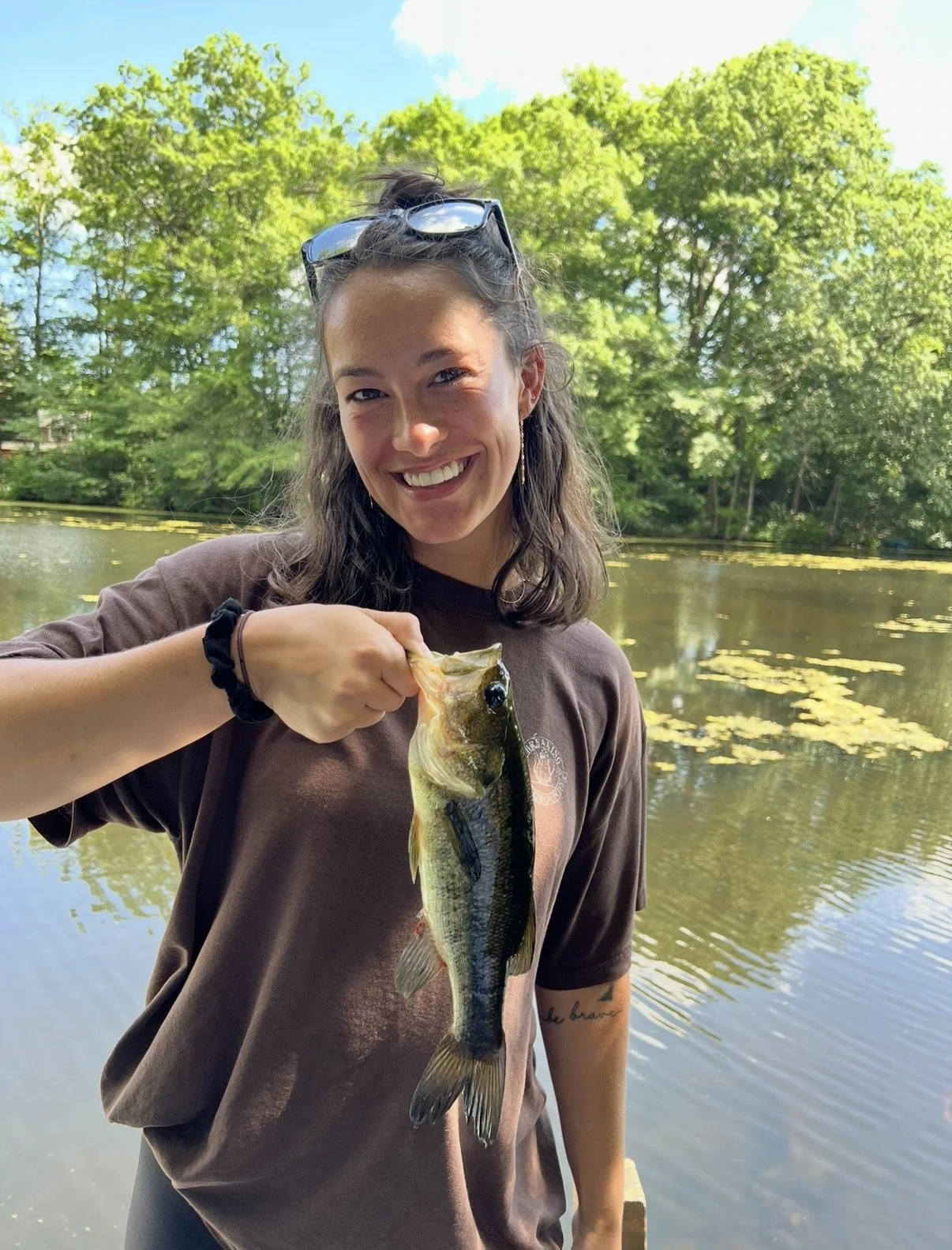 A woman smiling and holding a fish by the mouth near a pond surrounded by green trees on a sunny day.