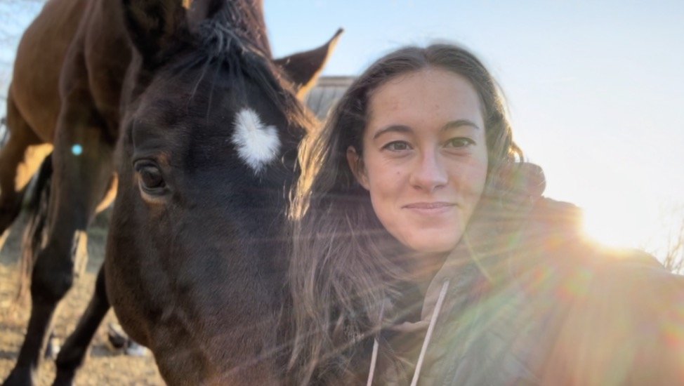 A young woman with long brown hair takes a selfie with a brown horse that has a white star on its forehead, outside during sunset or sunrise.
