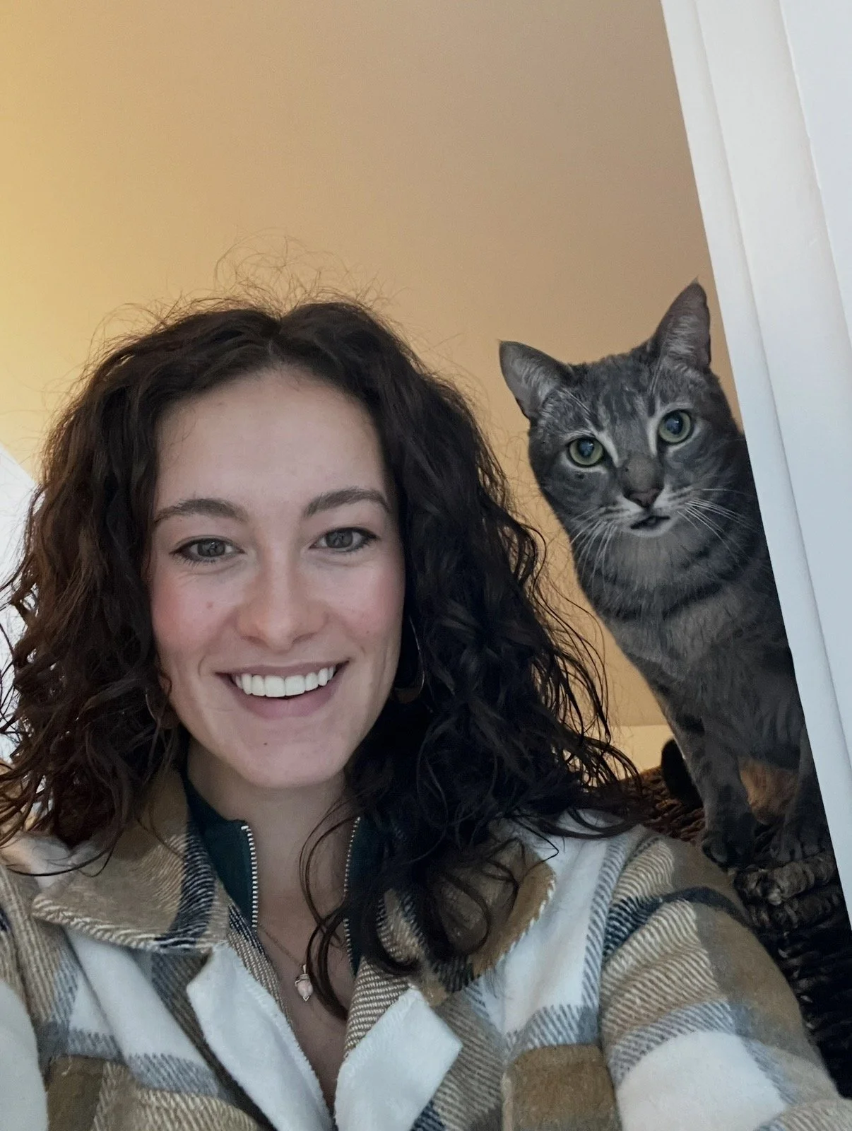 A woman with curly brown hair smiling and a gray tabby cat with green eyes peeking around a corner.