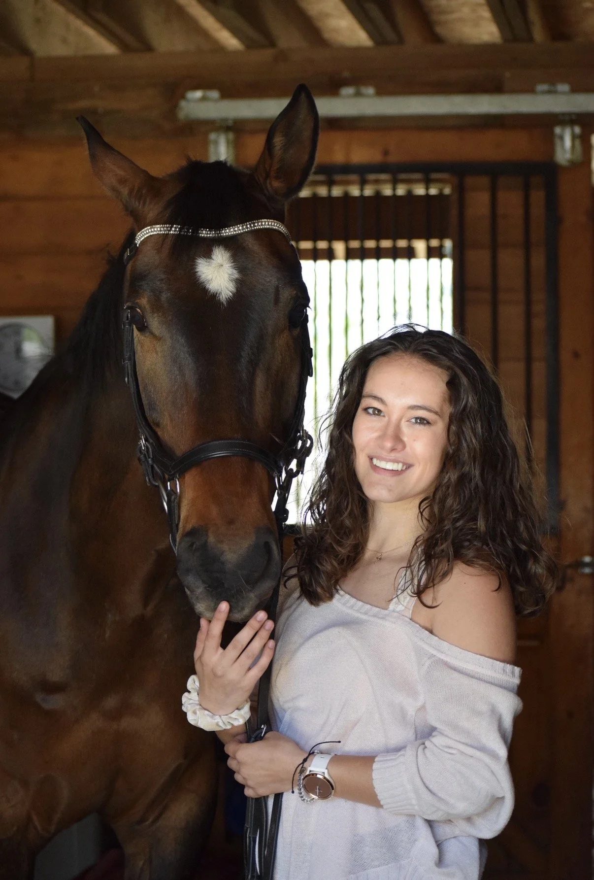 A young woman with curly brown hair and a white off-the-shoulder top smiling and holding a brown horse with a white star-shaped marking on its forehead inside a wooden stable.