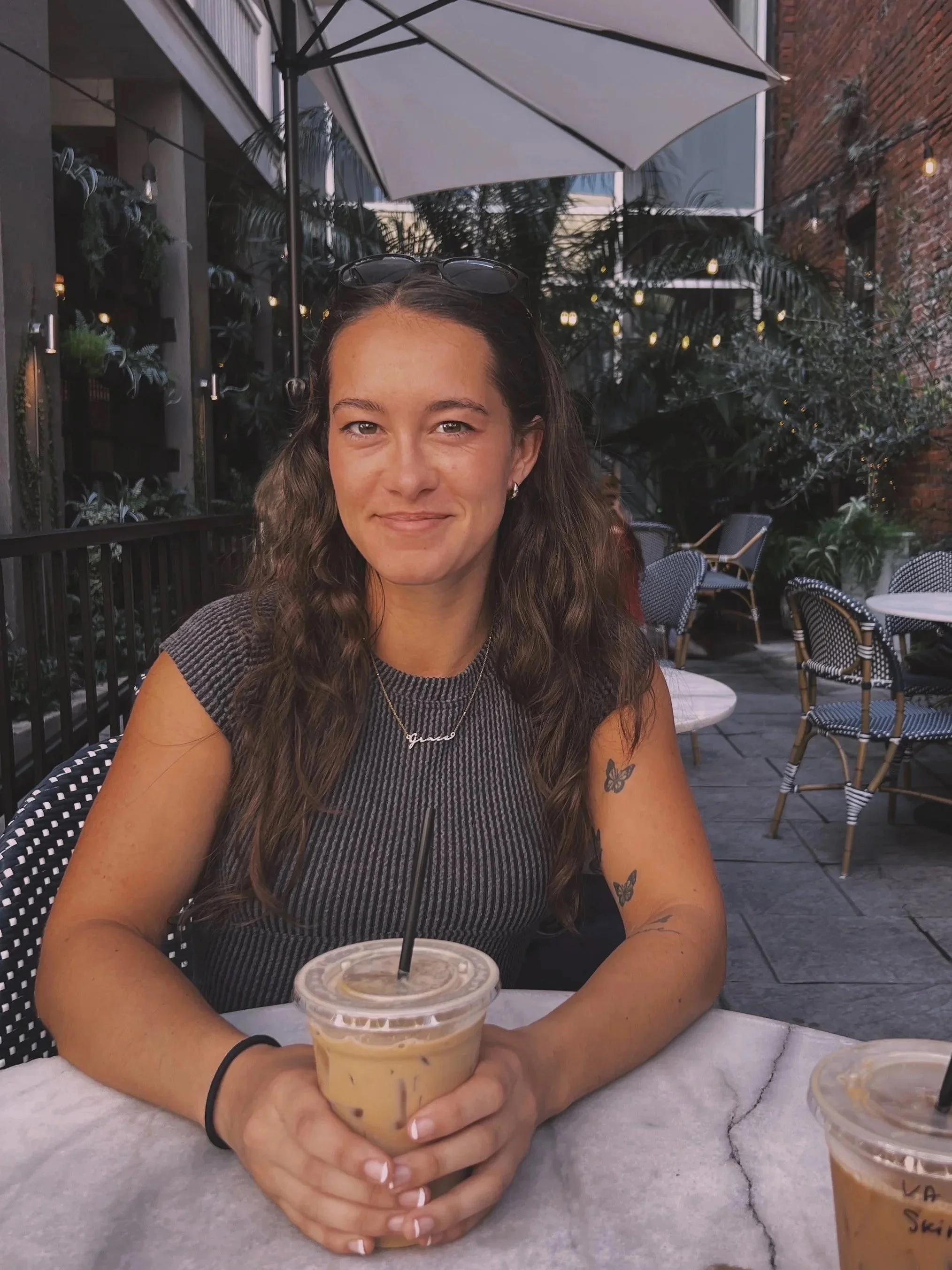 A woman with long curly brown hair, light skin, and tattoos of butterflies on her arm, sitting at an outdoor cafe holding a iced coffee. She is wearing a black bracelet, a necklace, and sunglasses on her head. The cafe has green plants, string lights, and tables with chairs around.
