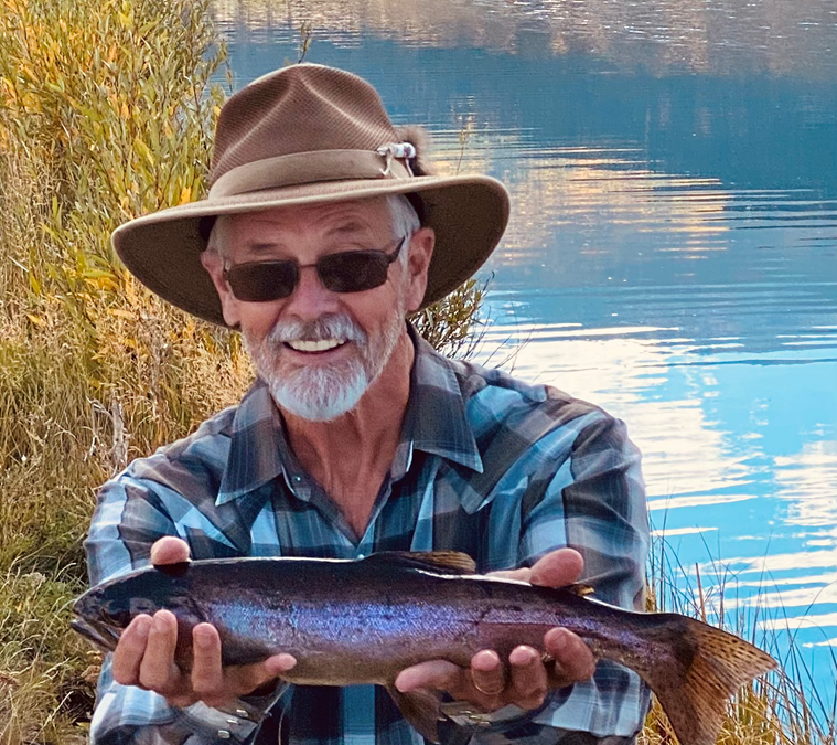 An older man with sunglasses, a wide-brimmed hat, a gray beard, and a checkered shirt holding a fish outdoors near a body of water with tall grass and blue sky.