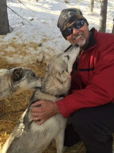 A man wearing sunglasses and a red jacket kneels outdoors in a snowy forest, hugging a Siberian Husky. Two other huskies are nearby, one licking the man's face.