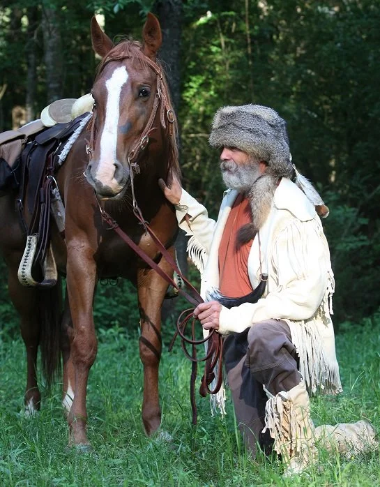 Older man in historical costume with fur hat, fringed coat, and boots, kneeling and holding the reins of a brown horse with a white blaze on its face in a forest clearing.