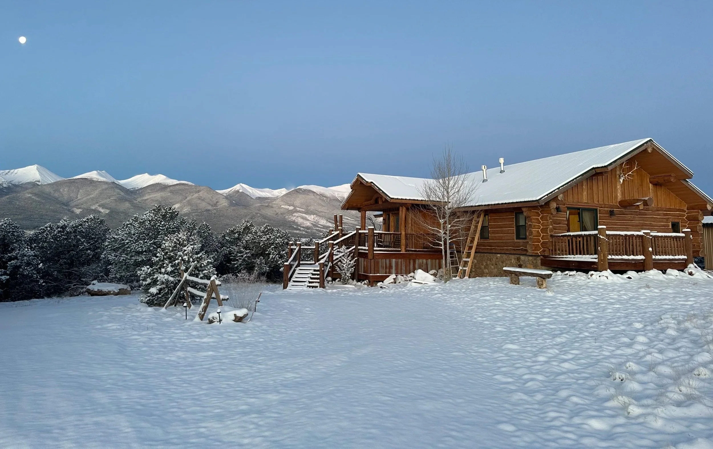 A snow-covered wooden house with a deck and stairs in a rural area with snow-covered trees and distant mountains under a clear blue sky with visible moon.