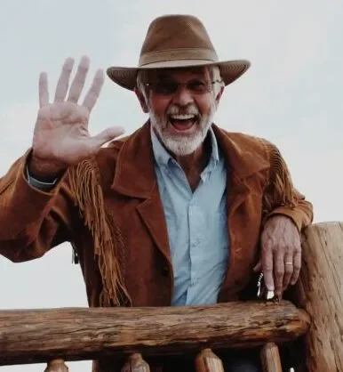 A smiling man wearing a cowboy hat, sunglasses, a brown jacket with fringe, and a light blue shirt, waving and leaning on a wooden railing outdoors.