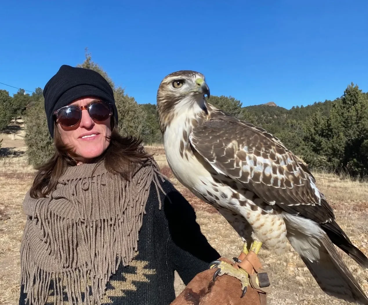 A woman wearing sunglasses, a black beanie, and a fringed scarf, holding a large bird of prey on her gloved arm in an outdoor setting with trees and mountains in the background.