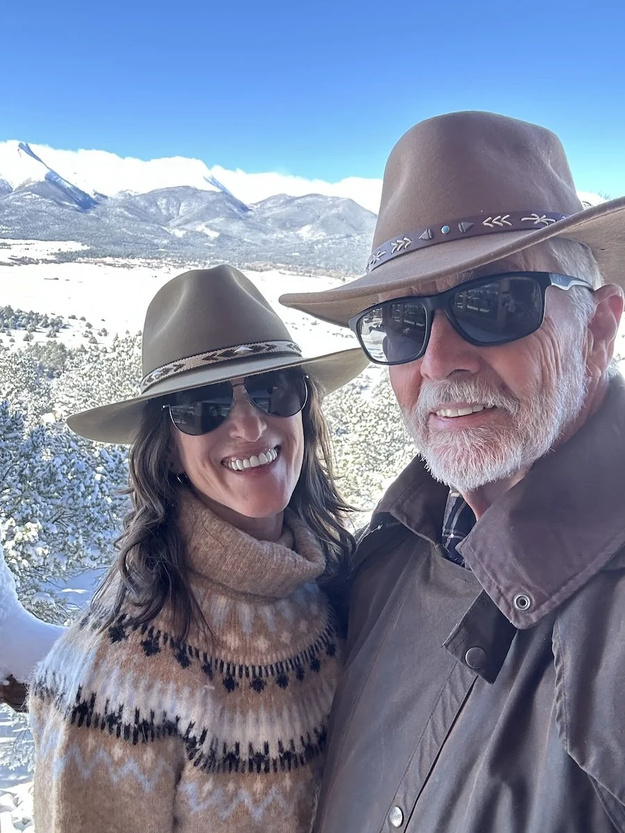 A smiling couple wearing hats and sunglasses taking a selfie outdoors in a snowy mountain landscape.