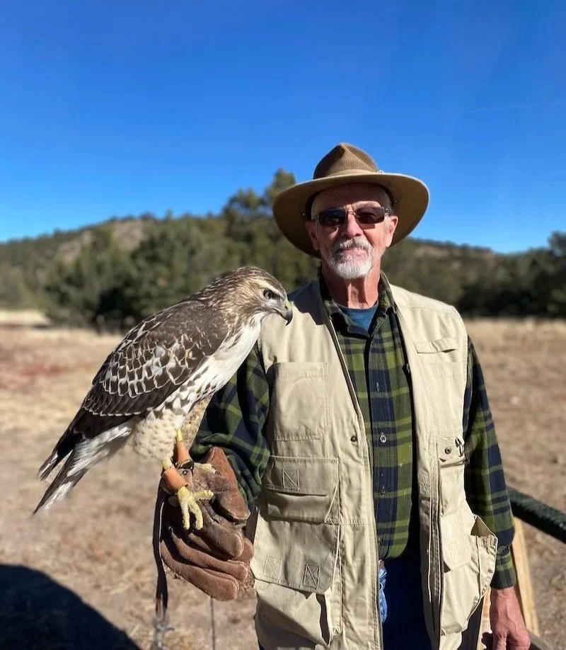 Older man wearing a wide-brimmed hat, sunglasses, and a beige vest holding a bird of prey on his gloved arm outdoors.
