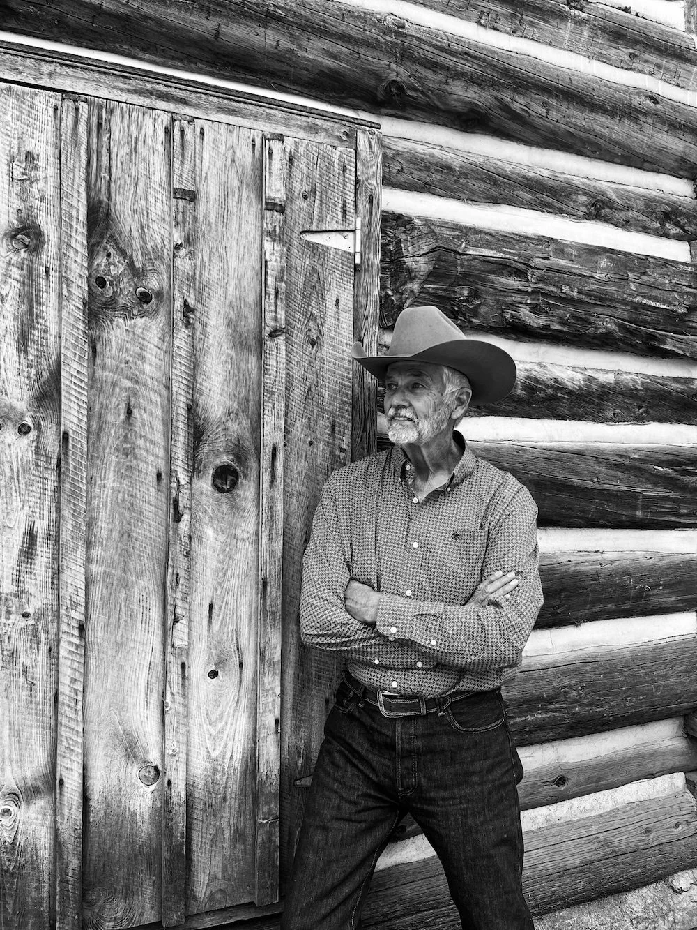 An elderly man with a beard wearing a cowboy hat, long sleeve shirt, and jeans standing against a wooden cabin wall with arms crossed.