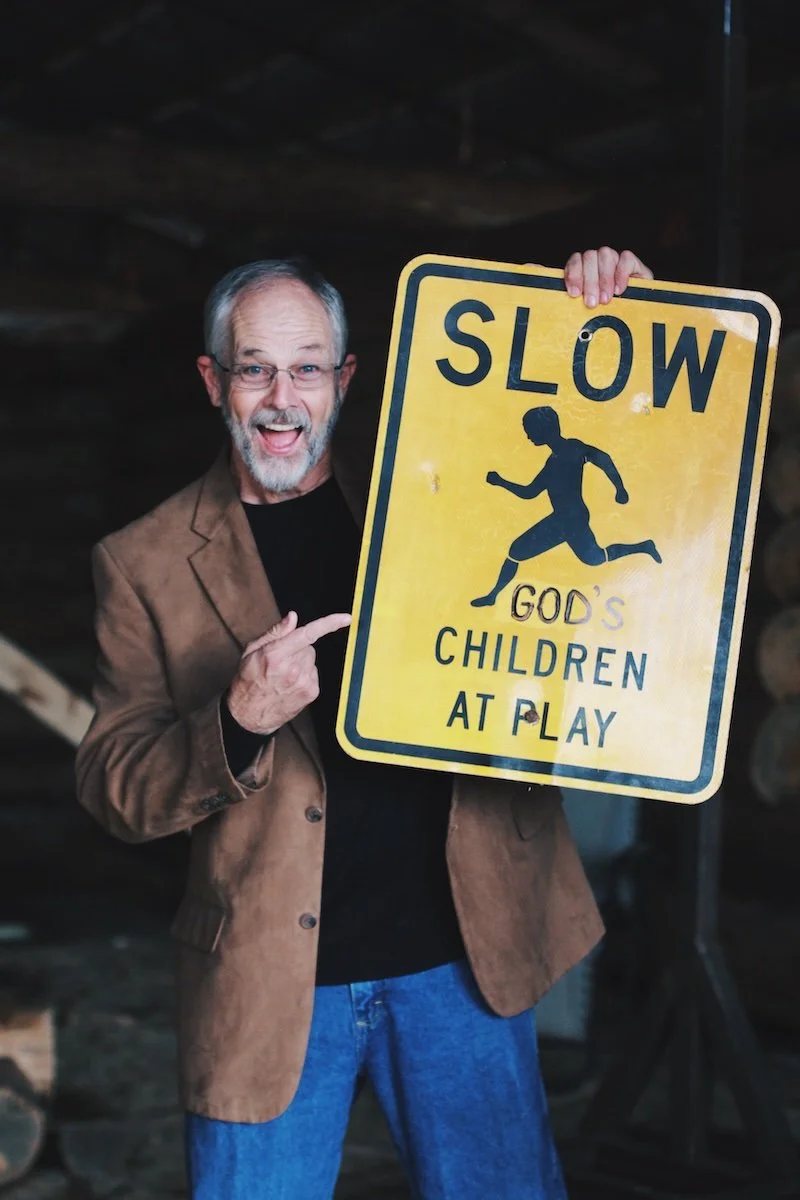 A man with gray hair and glasses wearing a brown blazer pointing at a yellow road sign that says 'SLOW' with an image of a running child and the words 'God's children at play' underneath.