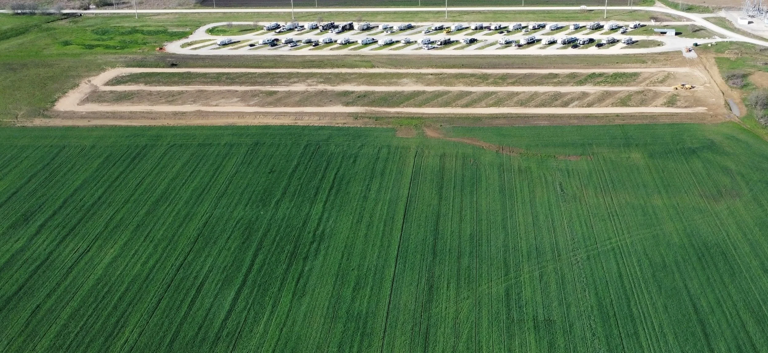 Aerial view of a construction site next to green fields, with several parked RVs and construction equipment in the background.
