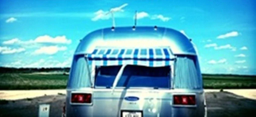 A silver vintage camper trailer with a blue and white striped awning, parked on a road with green fields and a blue sky with clouds in the background.