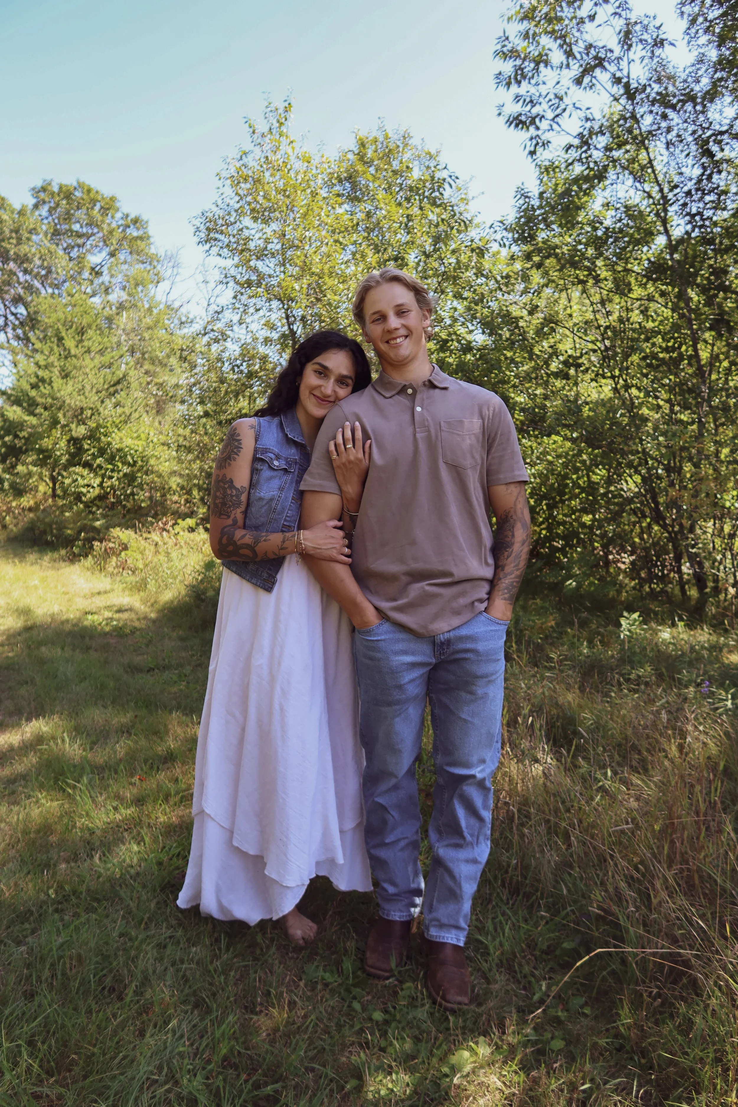 A smiling couple standing outdoors embraced, the woman wearing a white dress and a sleeveless denim jacket with tattoos on her arms, the man wearing a beige polo shirt and light blue jeans, with green trees and blue sky in the background.