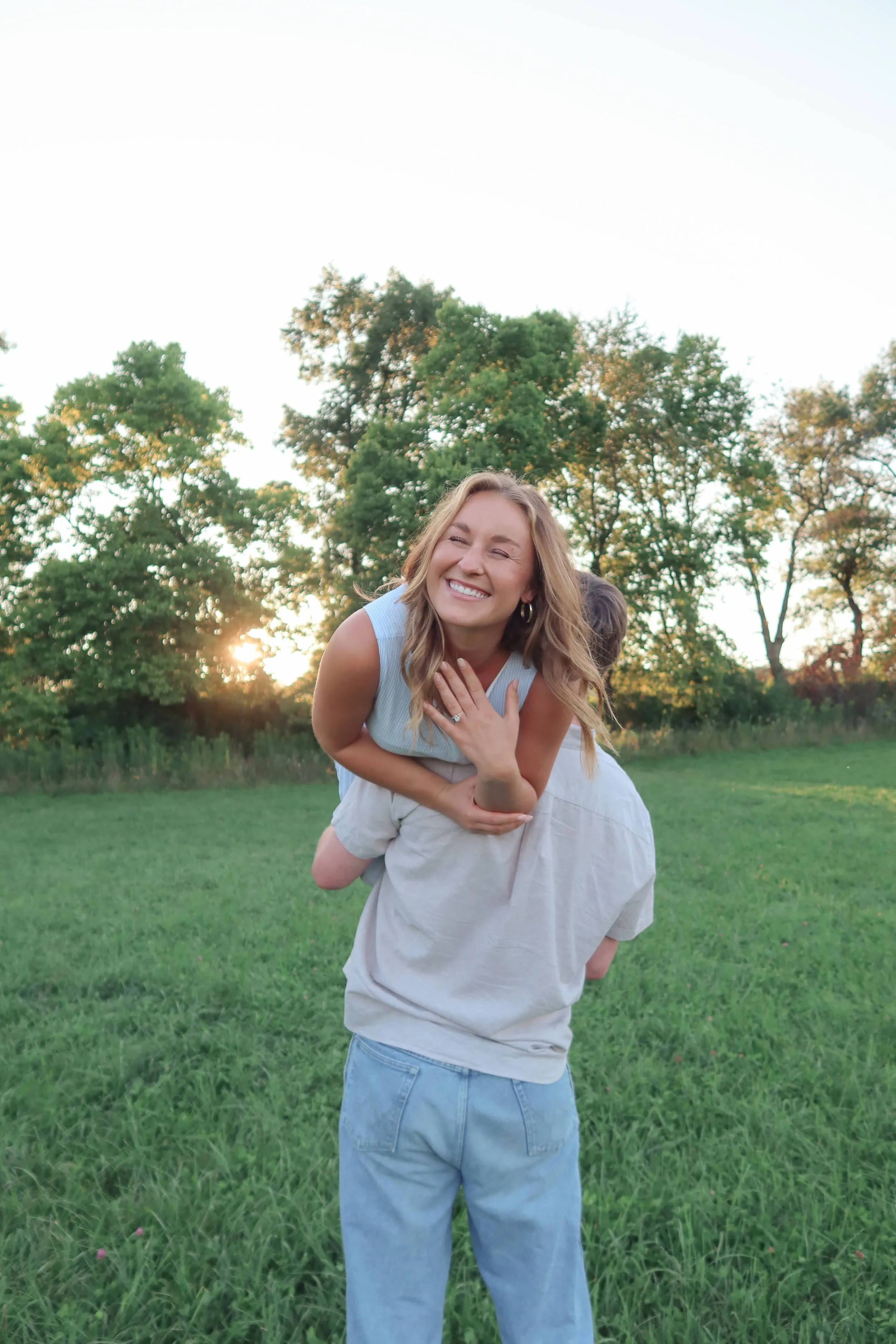 A woman with long, wavy blonde hair, smiling and laughing, with her hand over her chest, outdoors on a grassy field with trees in the background during sunset.