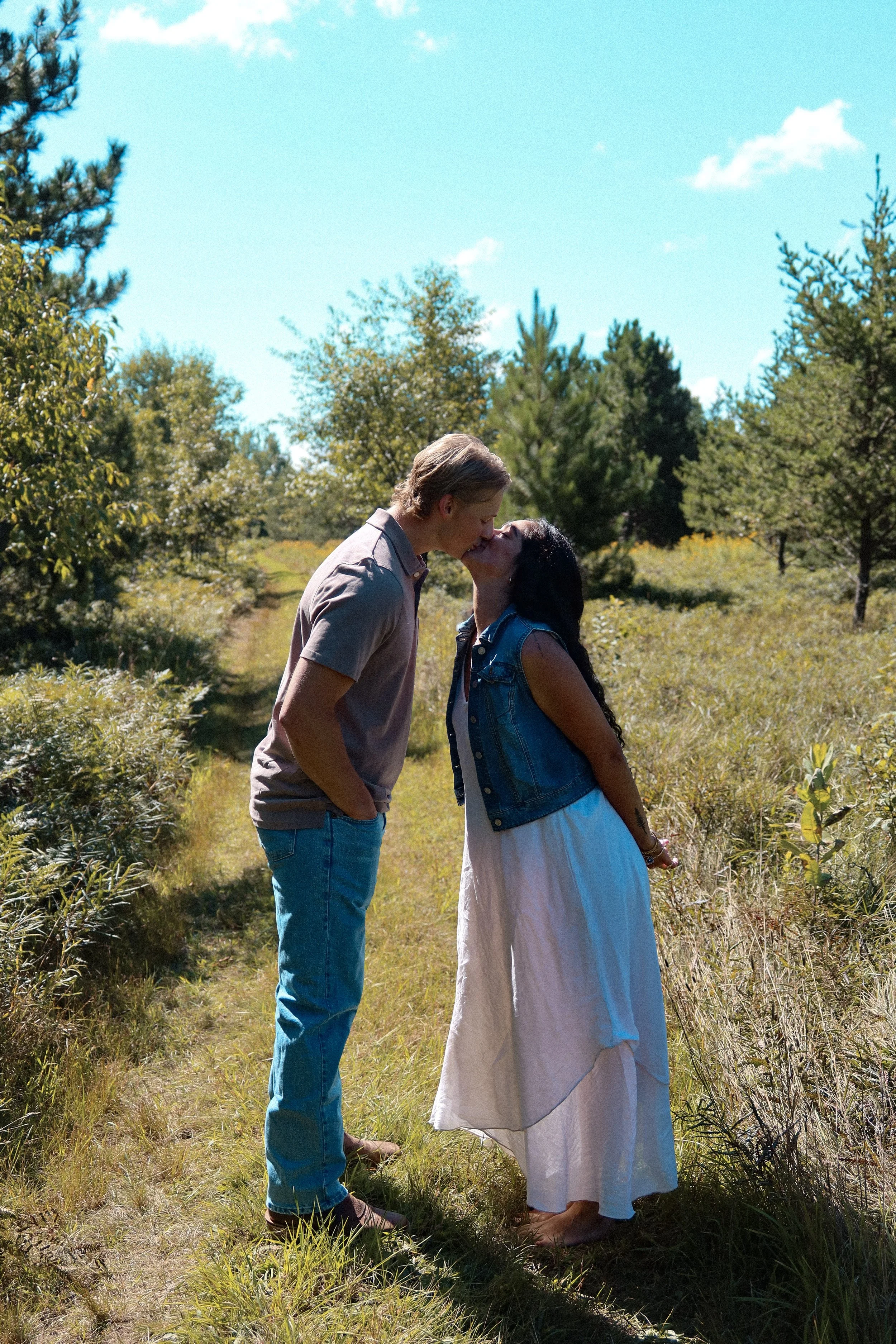 A couple stands in a grassy field surrounded by trees on a sunny day, kissing.