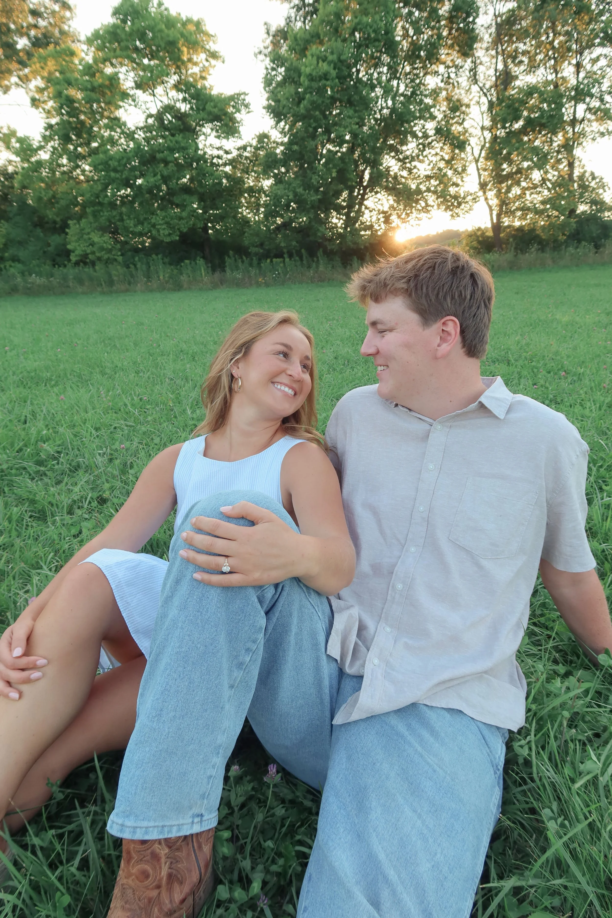 A smiling woman and man sitting on grass in a field at sunset, gazing at each other, with trees and a setting sun in the background.